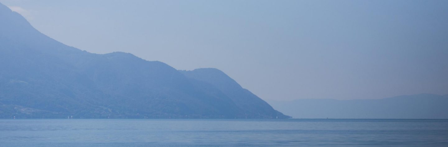 Ruhige blaue Seelandschaft mit sanften Wellen und dunstigen Bergen unter blassblauem Himmel.
