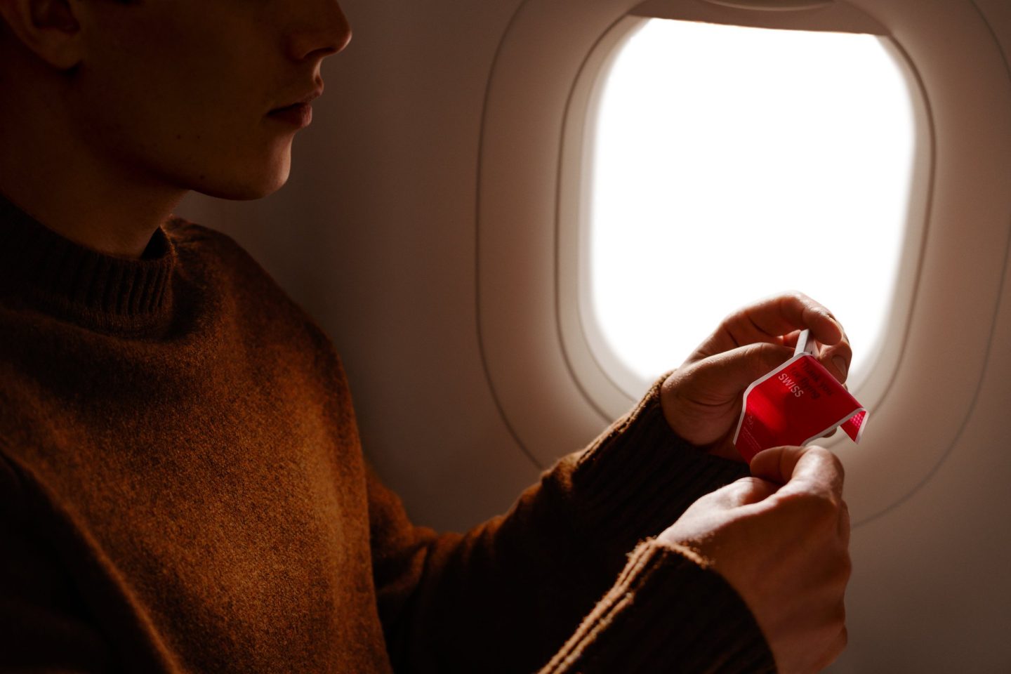 Passenger in a brown jumper by an aircraft window opening a red Swiss chocolate wrapper.