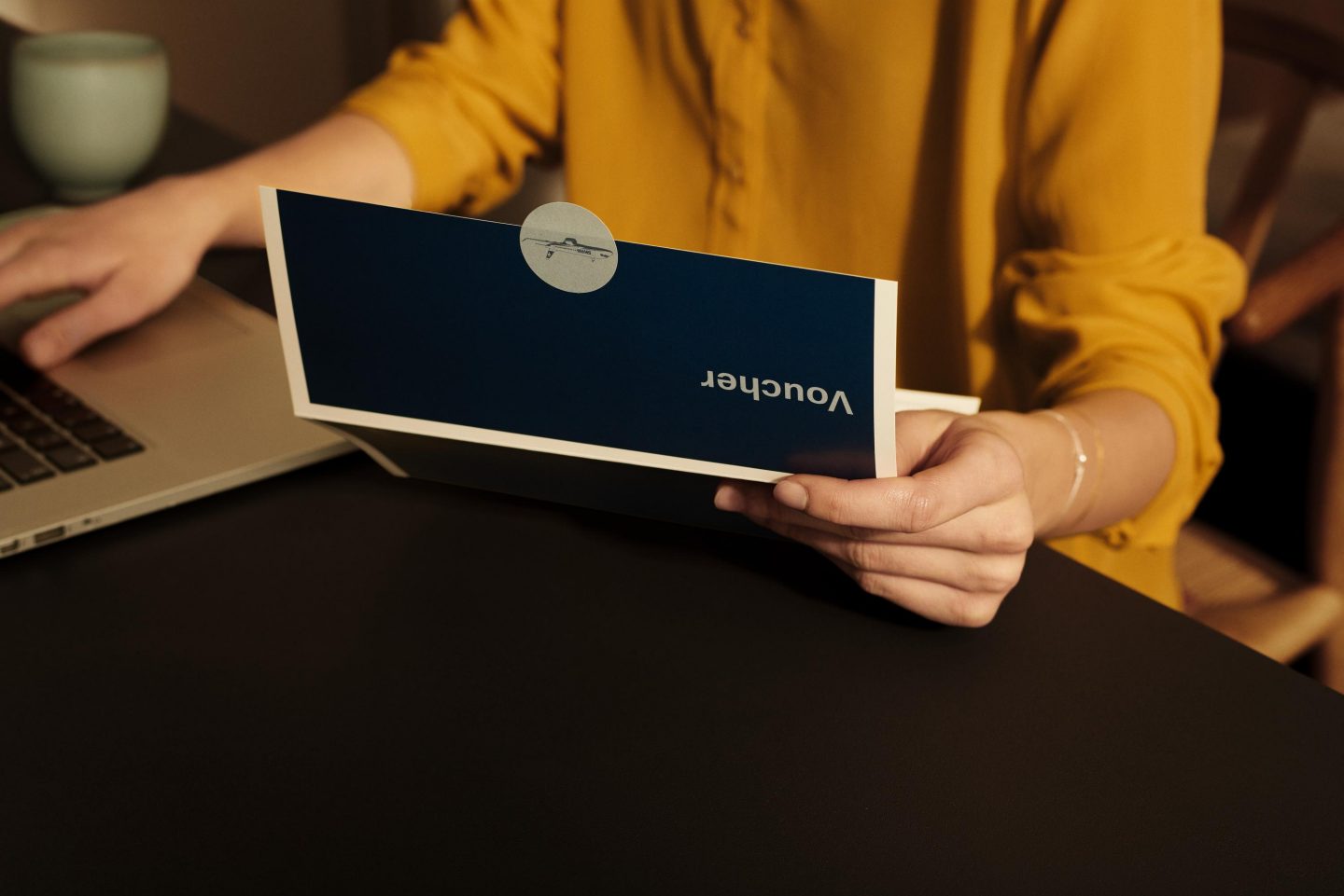 woman in yellow blouse at home in front of laptop holding a voucher in her hand