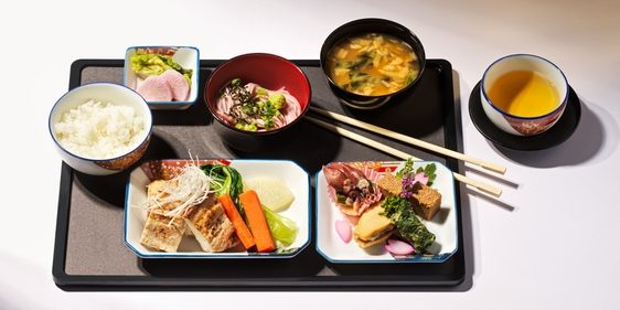 A tray with Japanese food on board an aircraft. The meal includes rice, a bowl of miso soup, various side dishes, vegetables and tea.