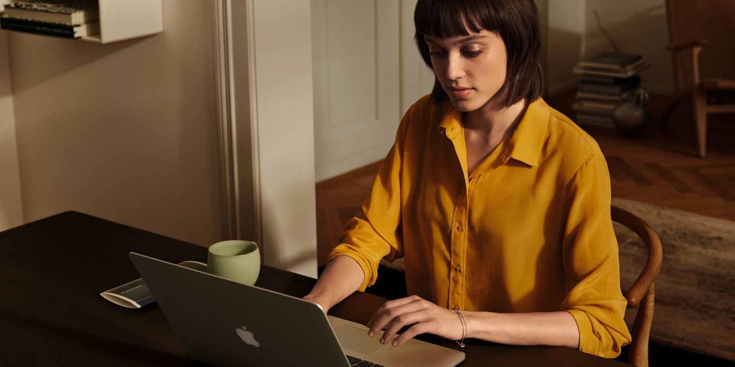 Une femme portant une chemise jaune assise à un bureau, travaillant sur un ordinateur portable.