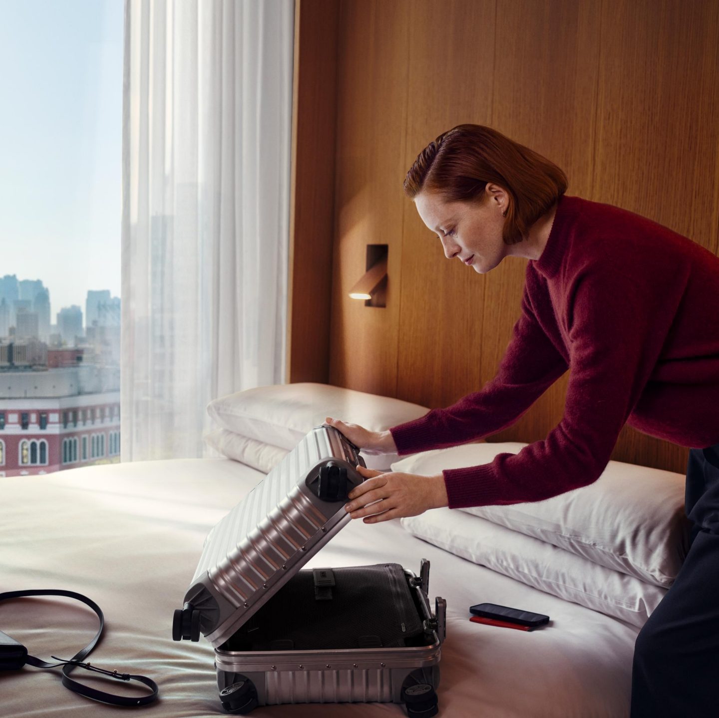 Eine Frau in einem roten Pullover packt auf einem Bett in einem Hotelzimmer einen silbernen Koffer aus, während durch das Fenster die Skyline der Stadt zu sehen ist.