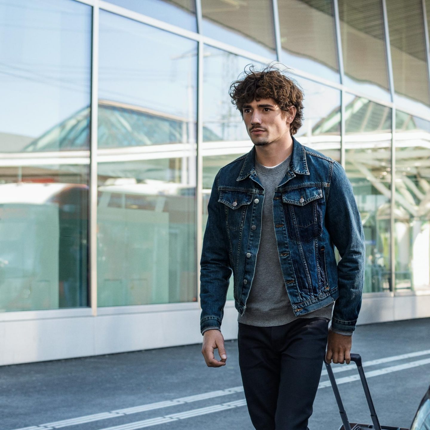 A young man walking with a suitcase outside a modern building.