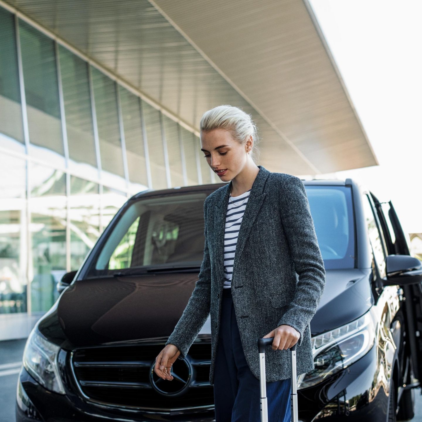 Blonde woman pulling a rolling suitcase in front of a luxury black van outside a terminal.