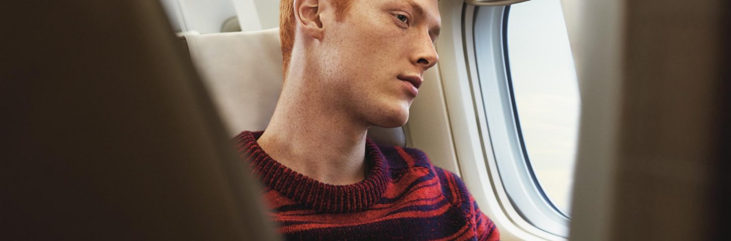 Rothaariger Passagier hält ein Smartphone und blickt aus dem Fenster einer SWISS-Flugzeugkabine.