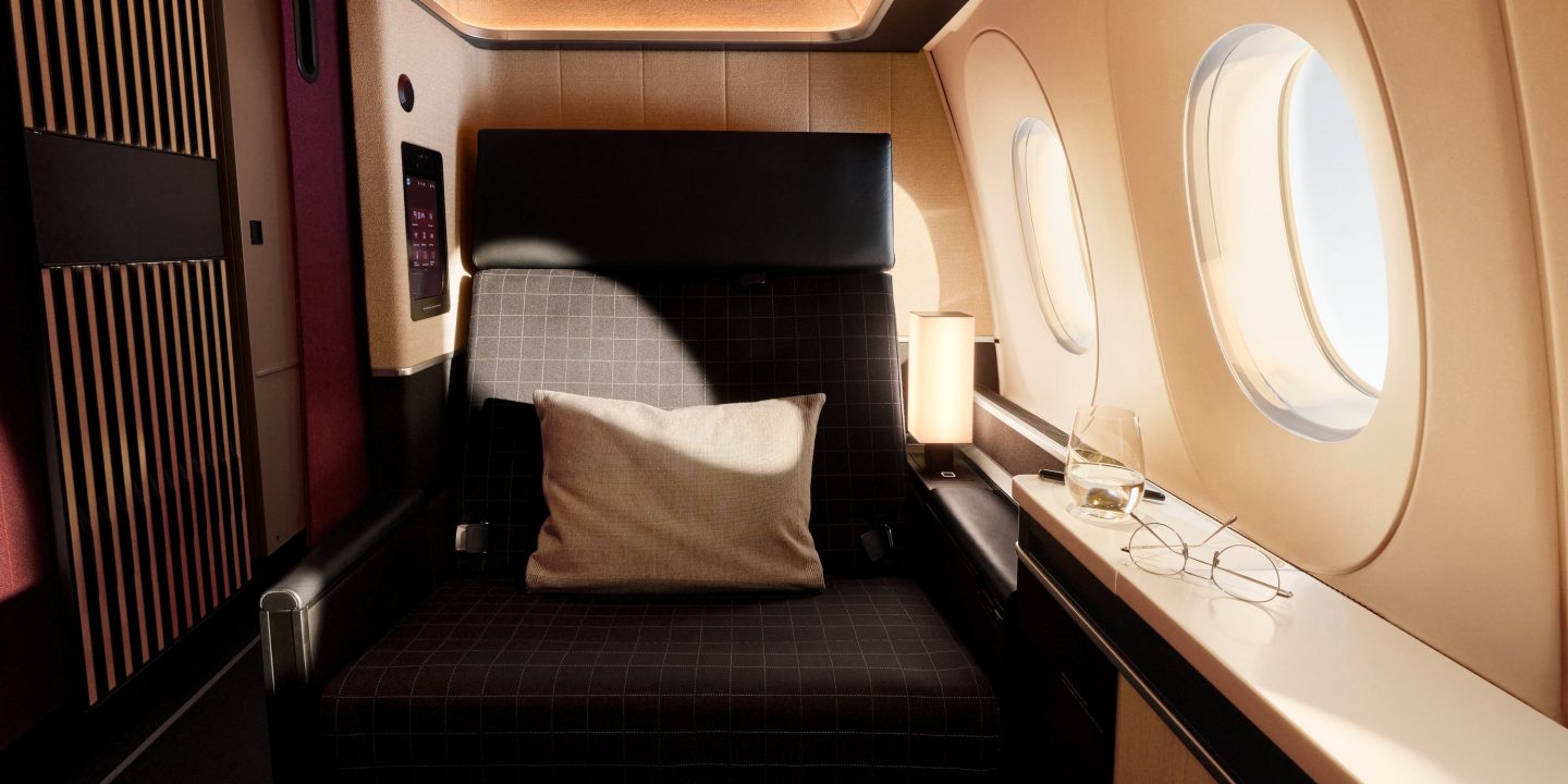 Luxury first-class airplane suite with a plush seat, side table, lamp, and windows.