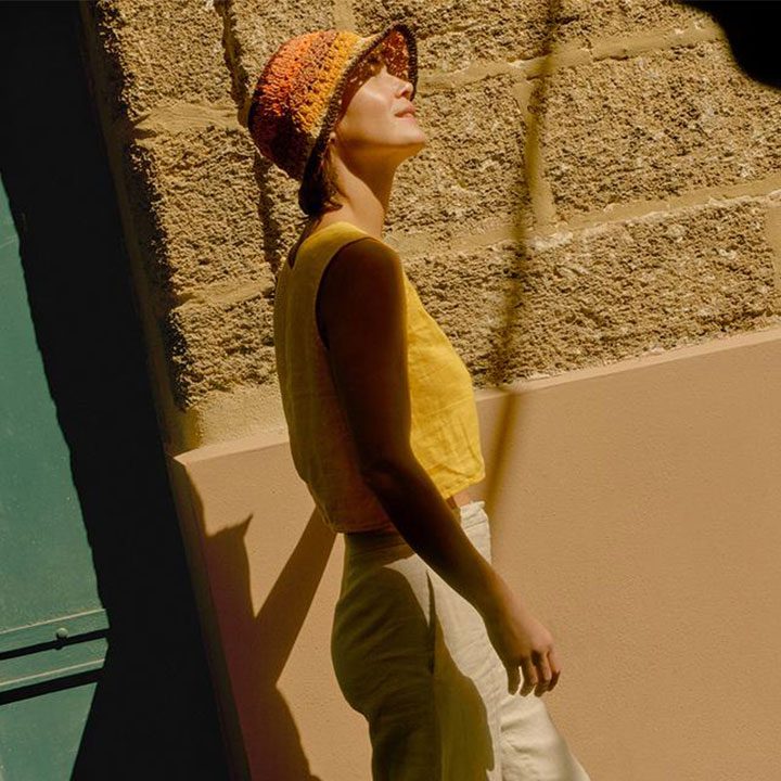 A woman in a yellow top and hat walking in sunlight beside a stone wall.