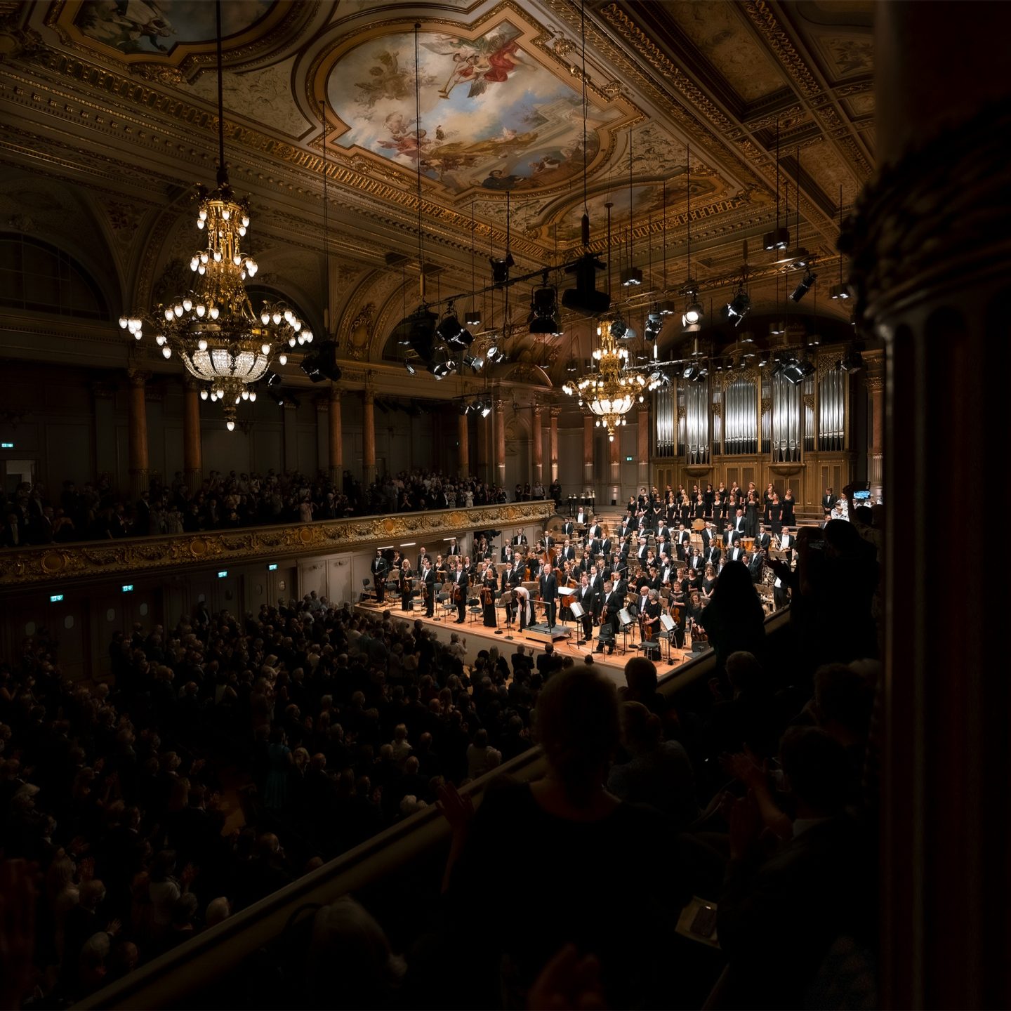 L'orchestra e il coro si inchinano sul palco di una grande sala da concerto con soffitto decorato e lampadari, mentre il pubblico applaude.