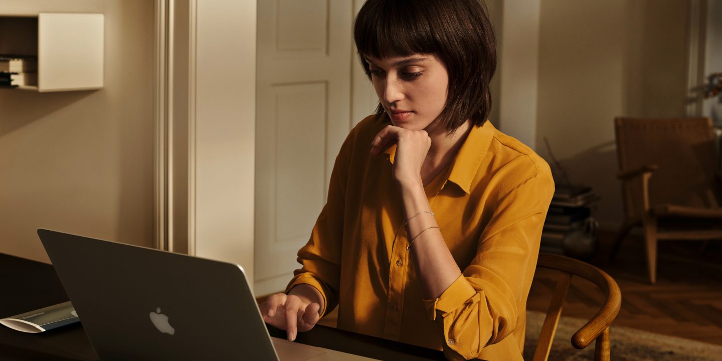 A woman in a yellow shirt uses a laptop at home.