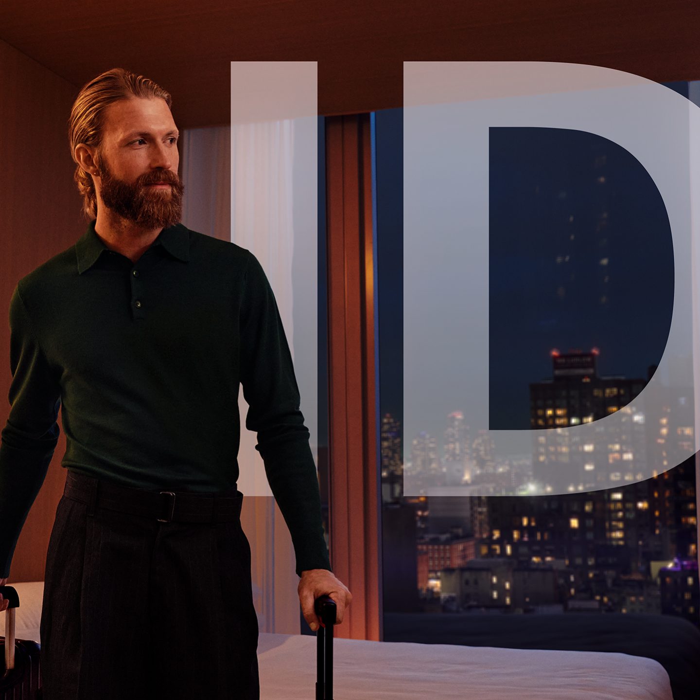 Bearded man with luggage in a modern hotel room overlooking a city at night.