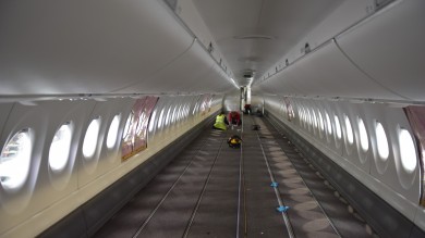 Der Flugzeuginnenraum ist zusehen. Nur ist dieser noch komplett leer, da noch keine Sitzreihen installiert wurden. Es sind drei Mechaniker:innen zu sehen, die die Vorbereitungen dafür treffen.