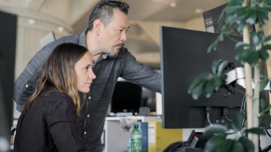 Standing up, a male colleague of Angela's shows her something on one of her many screens. Angela sits and listens attentively. 