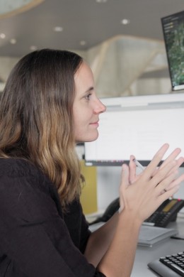 NOC manager Angela is seen in side profile, sitting in front of her desk. the desk is equipped with 4 screens, one of which is on top of the other three. it shows a map. 