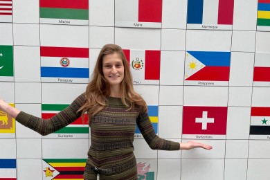 A woman stands in front of a wall showing many different country flags. 