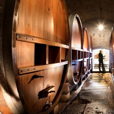 In einem Weinkeller befinden sich grosse Weinfässer. Hinten im Bild ist ein Mann ersichtlich. Der Weg zu ihm ist mit steinen gepflastert und Nass vom Wein.