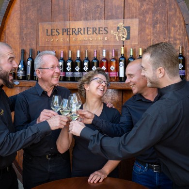 Abgebildet ist eine Familie, bestehend aus vier Männern und einer Frau. Alle stossen mit einem Glas Wein an und lachen. Im Hintergrund stehen viele verschiedene Weinflaschen und das Logo des Familienweinguts. 