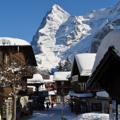 Personen flanieren durch die schneebedeckte Dorfstrasse. Der schneebedeckte Eiger ragt im Hintergrund empor.