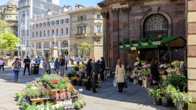 Der St. Ann's Square ist ein belebter Platz im Herzen von Manchester, umgeben von Geschäften, Kirchen und weiteren Sehenswürdigkeiten. Ein Blumenstand bringt lebendige Farben in den verkehrsberuhigten Raum.