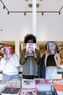 Three people explore UNITOM, holding magazines in front of their faces, immersed in the independent bookshop’s vibrant selection of visual arts publications.
