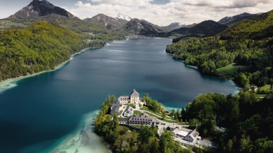 Ein atemberaubender Blick auf das Rosewood Schloss Fuschl, eingebettet auf einer ruhigen Halbinsel, umgeben vom kristallklaren Wasser des Fuschlsees und den majestätischen österreichischen Alpen.